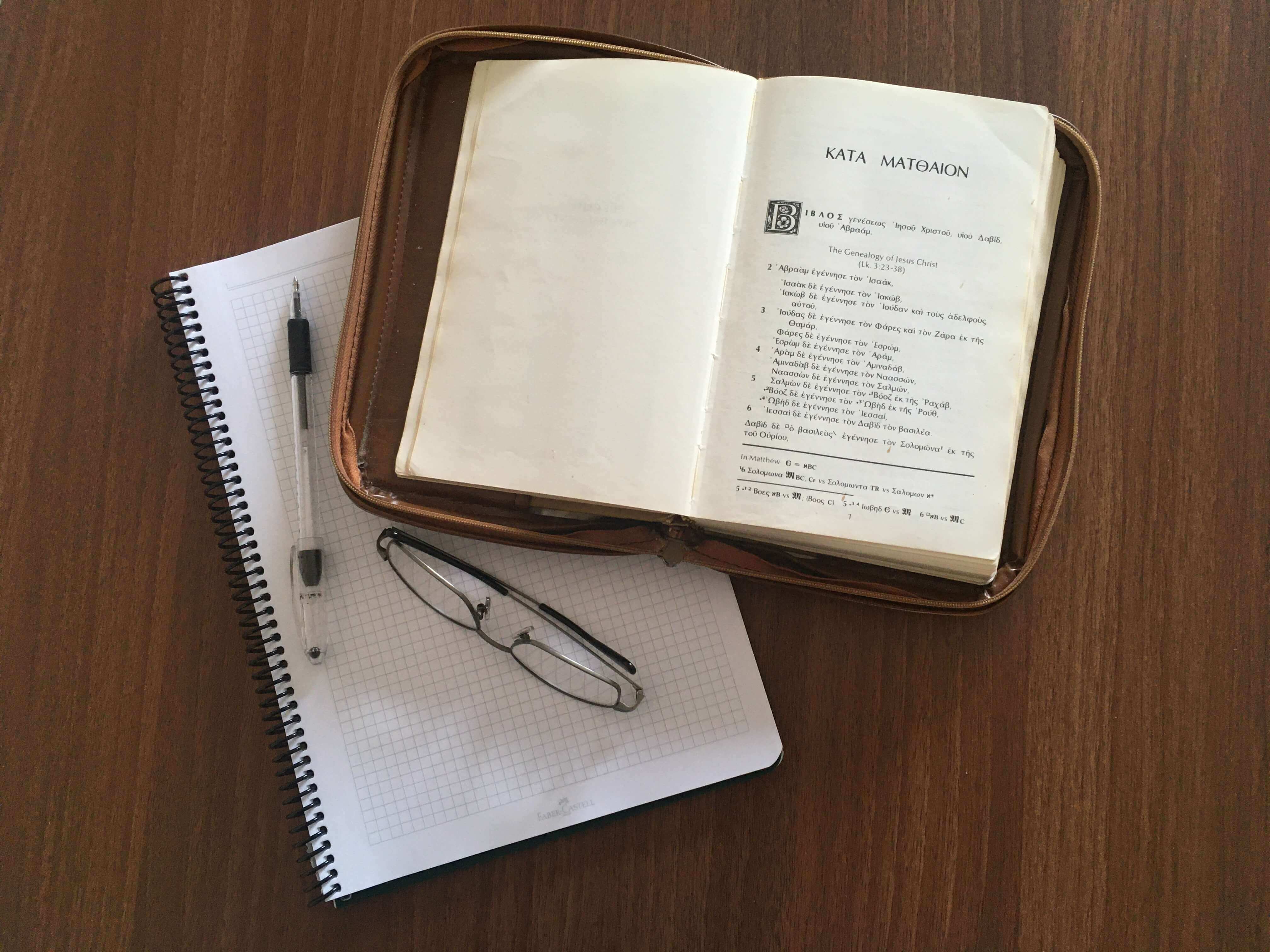 a notebook, a pen, a pair of glasses, and a Greek New Testament opened to Matthew 1